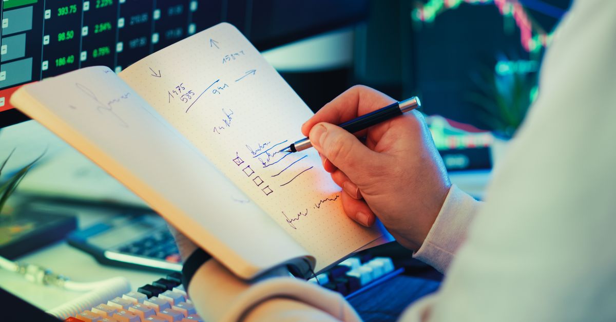 A trader writing notes by hand in a notebook while stock market data displays on screens in the background.