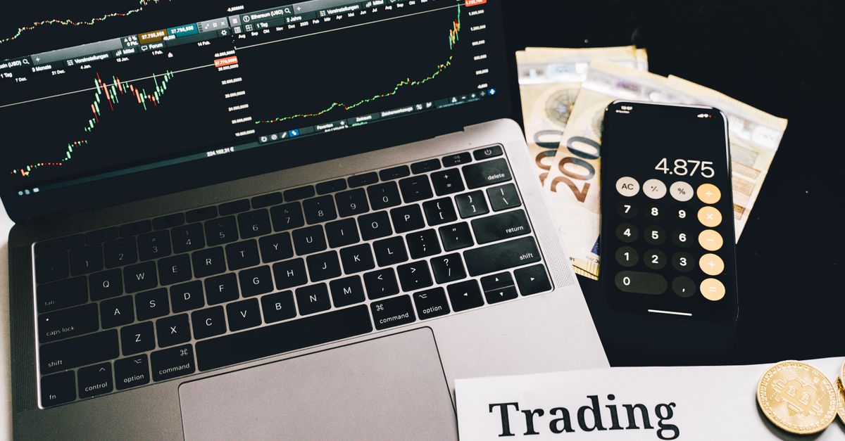 A laptop showing stock market graphs with a smartphone calculator, euro bills, coins, and trading strategies paper.