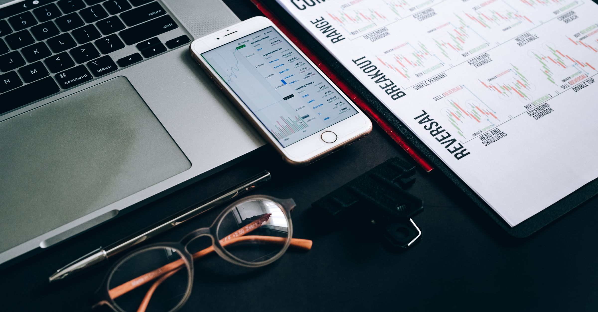Workstation with laptop, smartphone, eyeglasses, financial charts, and pen for trading analysis.