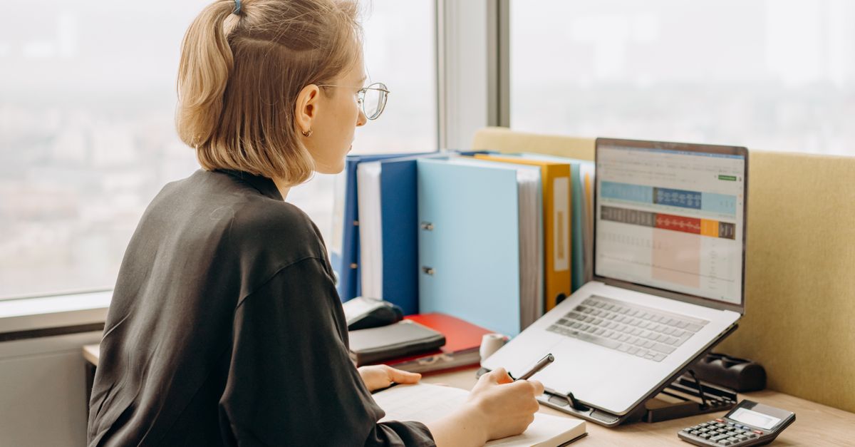 A trader reviewing a spreadsheet on a laptop at a desk with binders and a calculator nearby.